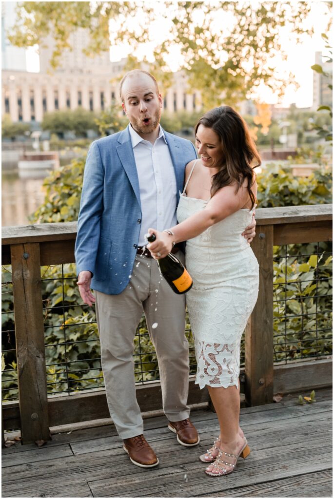 Engaged couple laugh as they pop a bottle of champagne. 