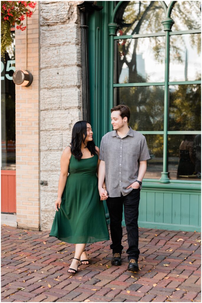 Engaged couple walking while looking at each other in front of large windows and brick.