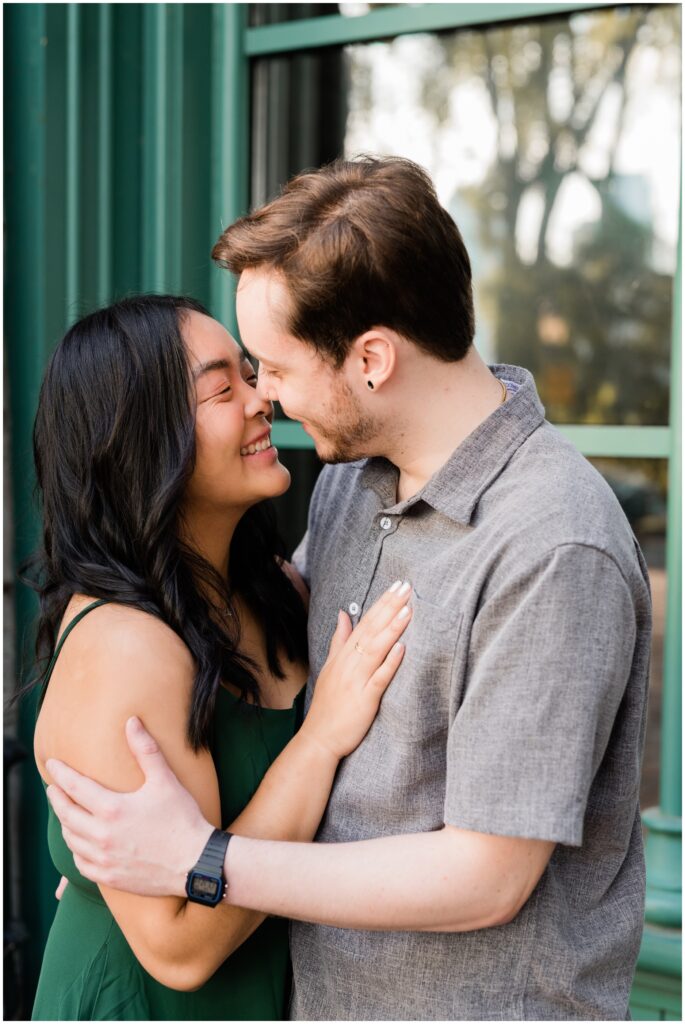 Engaged couple giggle kissing in front of windows.
