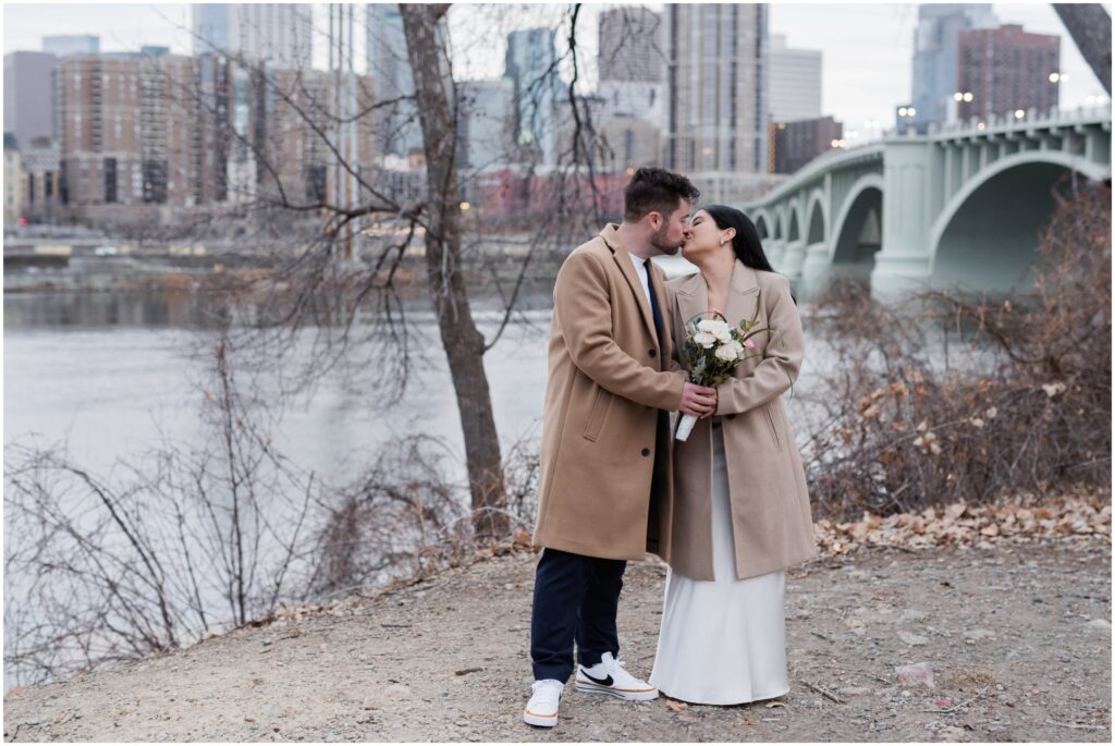 A bride and groom kiss with the skyline of Minneapolis in the background.