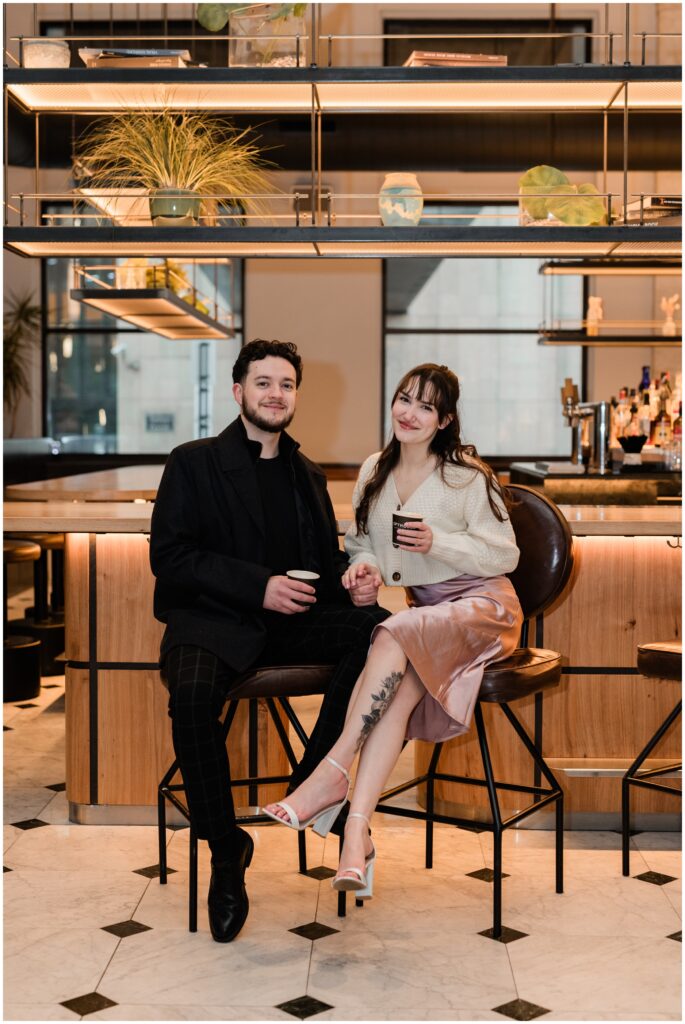 A couple sits at the bar while holding hands and holding coffees. 
